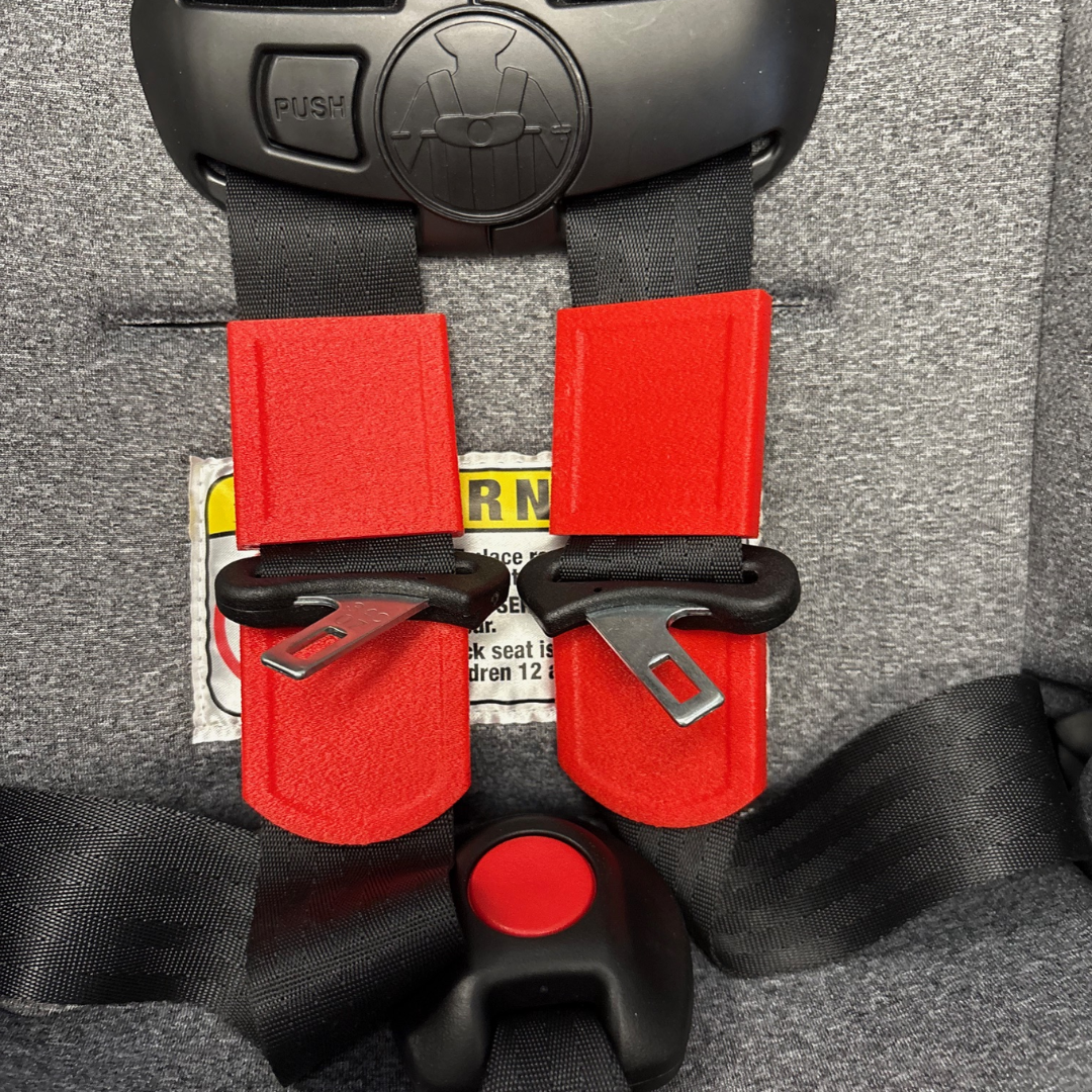 Close up photo of the red Buckle Buddeez device that keeps car seat straps from twisting installed on black car seat straps.