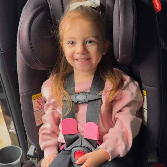 A smiling little girl in a car seat featuring pink BuckleBuddeez strap accessories.